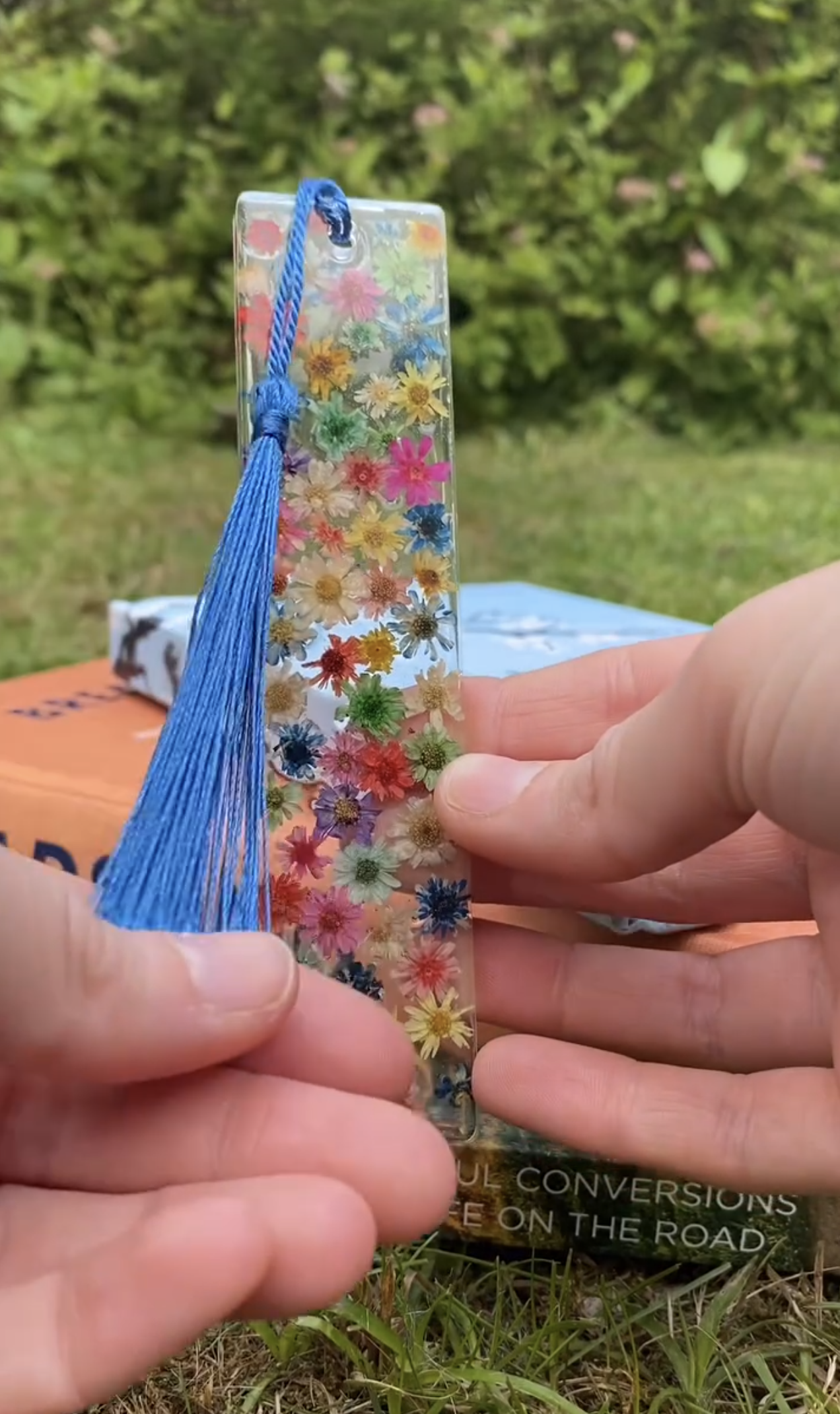 A colourful pressed-flower resin bookmark with a blue tassel, held in front of stacked books