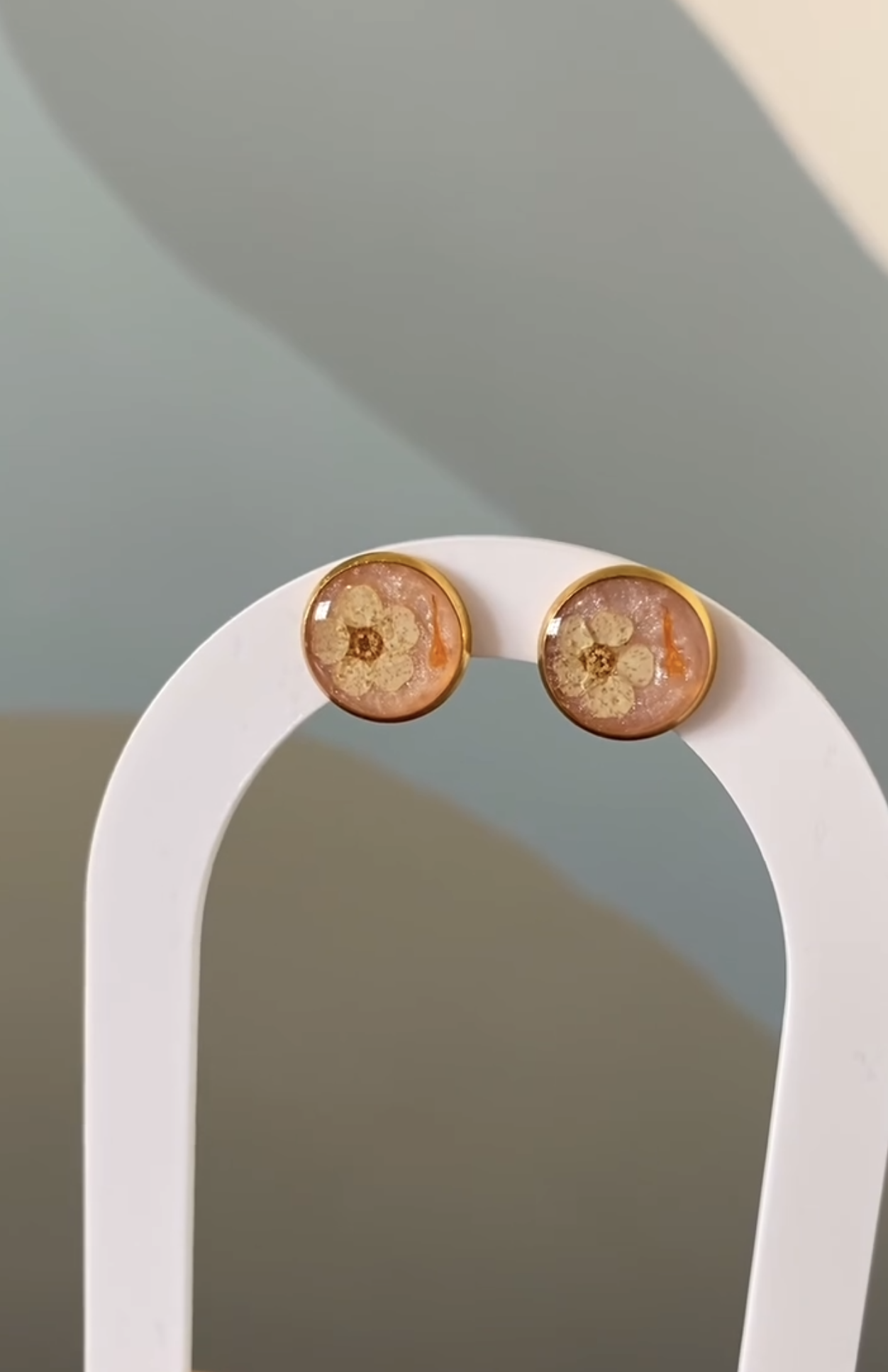A pair of gold-rimmed pressed flower stud earrings resting on a white arch jewellery display