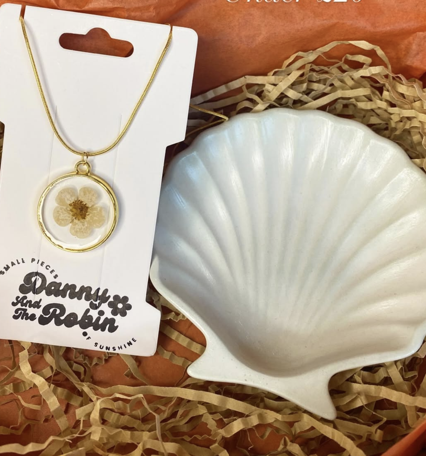 A pressed-flower gold necklace pendant alongside a white shell-shaped ceramic dish, displayed in an orange gift box